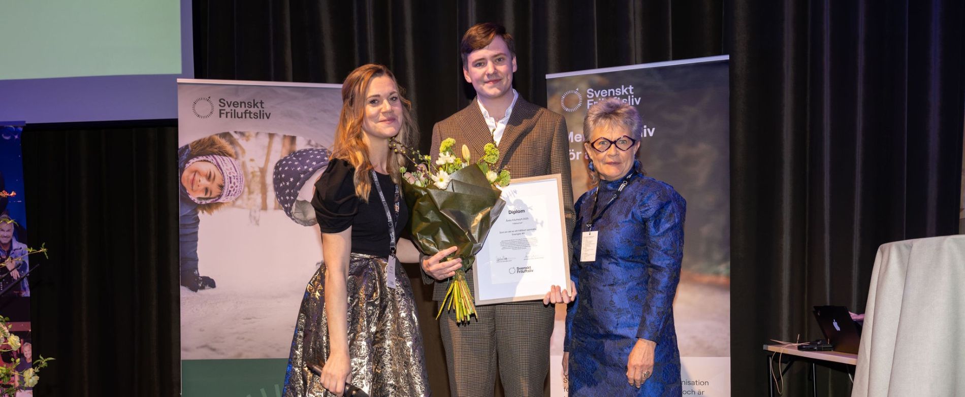 Bilden visar på tre personer som visar upp blommor och diplom i samband med utmärkelsen Årets friluftslyft 2025 på Svenskt Friluftslivs medlemskonferens.